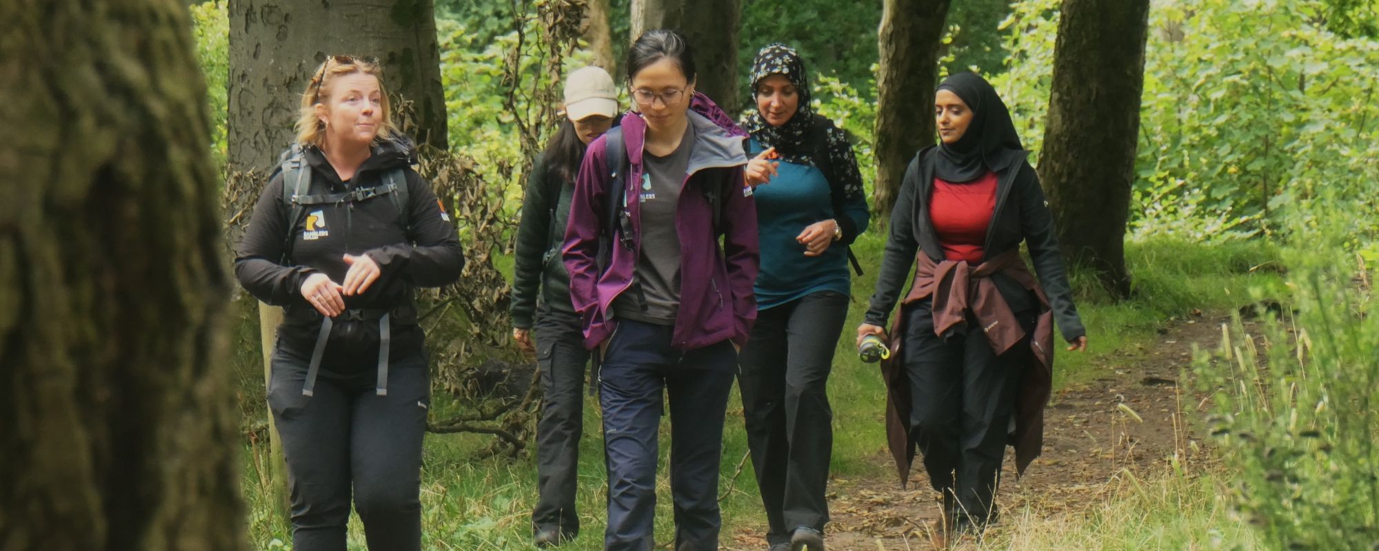 A group of five people is walking along a woodland trail surrounded by tall trees and green foliage. The people are dressed in outdoor clothing, including jackets, hoodies, and backpacks. They walk in a loose formation, with one woman in the foreground on the left and the others following slightly behind. The path is narrow with grass, soil, and roots visible underfoot. Sunlight filters through the trees, creating dappled light on the ground and surrounding vegetation.